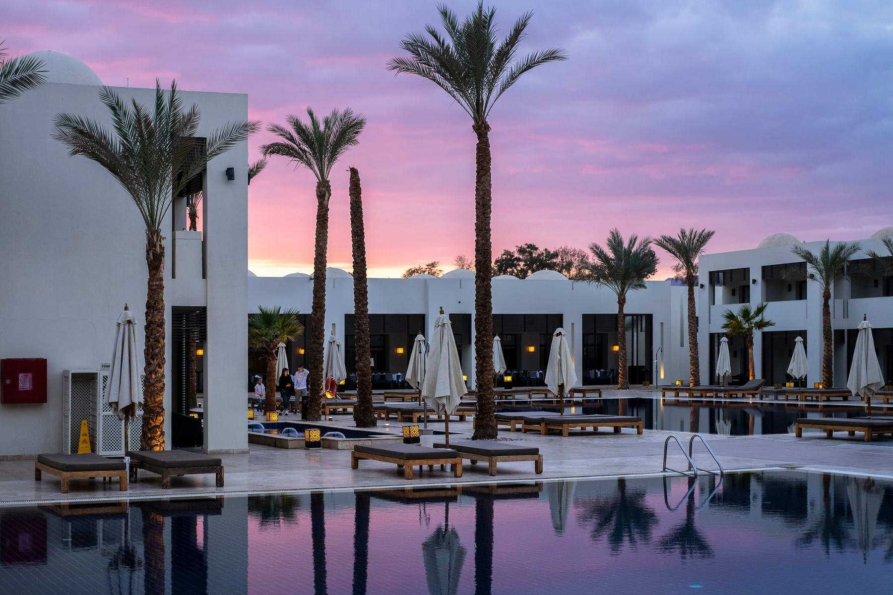 A white-walled resort at sunset, palm trees and sun loungers around a still pool reflecting a pink sky